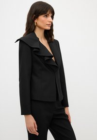 Blazer nero con un ampio colletto a ruches, chiusura a singolo bottone, maniche lunghe e vestibilità su misura, realizzato in un tessuto a trama liscia.