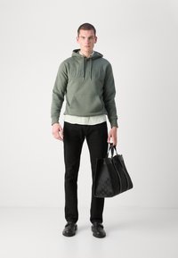 Sudadera verde con logo en relieve, superpuesta a una camisa clara. Pantalones negros, bolso negro con patrón geométrico y zapatos negros.
