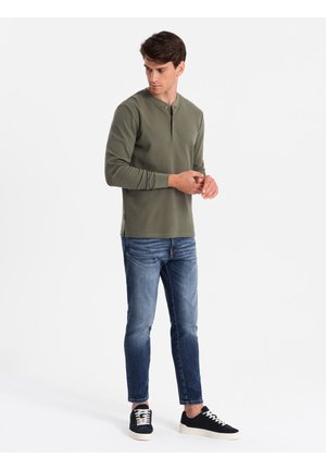 Maglietta henley a maniche lunghe verde oliva con scollo rotondo, due bottoni al collo, abbinata a jeans blu e sneaker navy.