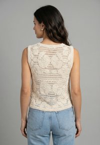 Mujer con cabello oscuro que lleva pendientes de perlas, top de crochet sin mangas color crema y vaqueros de mezclilla azul claro, vista desde atrás sobre fondo gris.