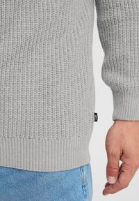 Grå teksturert strikkegenser med ribbestrikkede mansjetter og i nederkant, med en svart merkelappdetalj på siden, kombinert med blå denimjeans.