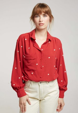 Blouse rouge à manches longues avec patte de boutonnage, poche et broderies florales blanches sur un tissu texturé. Accompagnée de pantalons couleur crème.