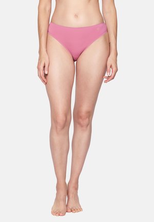 Frau steht barfuß in pinker nahtloser Bikini-Unterwäsche vor einem schlichten hellen Hintergrund.