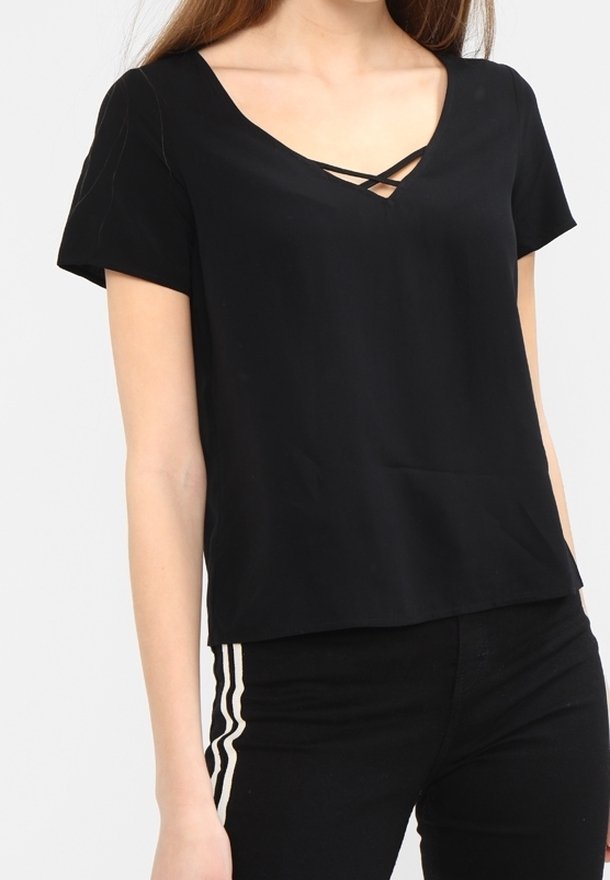 Femme portant un haut noir à manches courtes avec un col en V et un détail croisé, associé à un pantalon noir avec des bandes blanches sur les côtés.