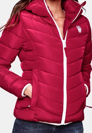 Femme portant une veste rouge matelassée avec une fermeture éclair blanche et un logo, les mains dans les poches, associée à un jean bleu foncé.