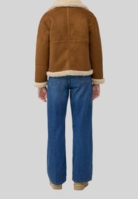 Bruine suède shearling jas met een kraag en manchetten van lichtbruine nepbont, gecombineerd met blauwe jeans met brede pijpen en beige laarzen, gezien van achteren.