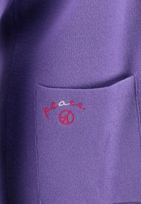 Paars stof met een zachte textuur, voorzien van een gestikte 'peace'-logo in rood en wit. Het ontwerp bevat een rond vredessymbool.
