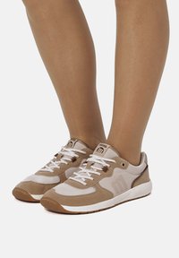 Beige Sneaker mit weißen und hellbraunen Akzenten. Merkmale sind ein Obermaterial aus Wildleder und Mesh, eine Schnürung und eine Gummisohle.