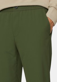 Pantaloni elastici verdi con una texture liscia, dotati di una tasca frontale e chiusura a bottone. La cintura è progettata per il massimo comfort.