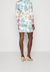 Femme portant une mini robe blanche ajustée avec des motifs floraux bleus, roses et verts, se tenant avec des talons beiges sur un fond uni.