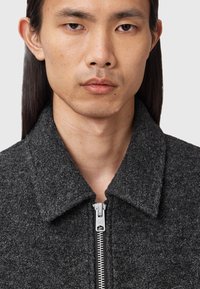 Grijze wollen jas met een opvallende kraag, voorzien van een zilveren metalen rits en een gestructureerd oppervlak. Close-up weergave richt zich op het bovenste kledingstuk.