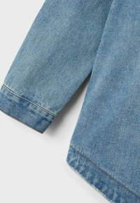 Lichtblauw denimjack met een recht gesneden mouw, rafelige zoom en zichtbare stikseldetails. Zachte textuur en een gladde afwerking.