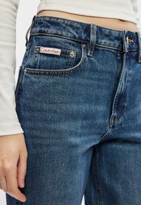 Jeansuri din denim albastru au un design clasic cu cinci buzunare. O etichetă vizibilă „Calvin Klein” este coasă pe buzunarul din spate, având accente din metal argintiu.
