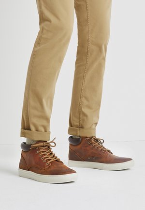 Braune Leder-Hochtop-Sneaker mit beigen Schnürsenkeln, cremefarbener Sohle und einem dunkelbraunen Akzent an der Ferse. Getragen mit hochgerollten Khaki-Hosen.