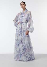 Mujer de pie lleva un vestido largo, translúcido, de flores en tonos pastel lila y azul, con mangas abullonadas y detalle de lazo al cuello, sobre un fondo liso.