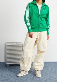 Zelená zipová bunda s bílými pruhy, kombinovaná s oversized krémovými kalhotami a béžovými teniskami. Jednoduchý design s minimem značkových detailů.