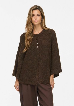 Frau mit langen hellbraunen Haaren, die eine lockere dunkelbraune flauschige Strickjacke mit vier Knöpfen und passende braune, weite Hosen trägt.