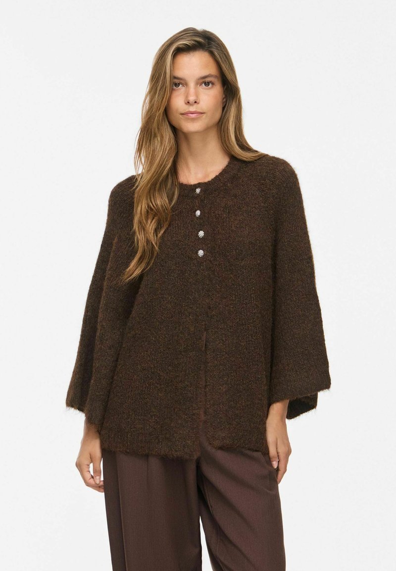 Femme aux longs cheveux châtain clair portant un cardigan duveteux marron foncé ample avec quatre boutons et un pantalon large marron assorti.