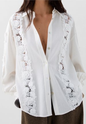 Chemise blanche boutonnée avec des détails en dentelle florale complexe le long des bords avant, tissu doux et coupe décontractée avec manches longues.