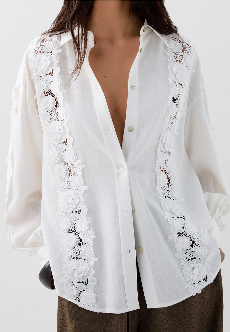 Chemise blanche boutonnée avec des détails en dentelle florale complexe le long des bords avant, tissu doux et coupe décontractée avec manches longues.