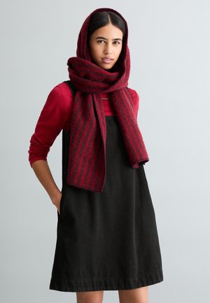 Sciarpa a righe rosse e nere drappeggiata su una maglia rossa a maniche lunghe e un vestito nero senza maniche. Il tessuto sembra morbido con cuciture e tasche visibili.