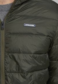 Padded jakna u tamnozelenoj boji s patentnim zatvaračem, kapuljačom i malim crno-bijelim logotipom "JACK & JONES" na prsima. Glatka tekstura.