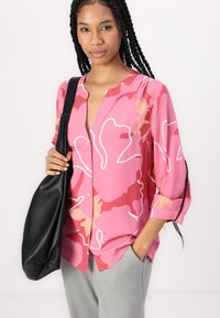 Bluse mit floralem Muster in Pink, V-Ausschnitt und Knopfverschluss. Mit hochgerollten Dreiviertelärmeln und grauen Hosen kombiniert.