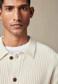 Beżowy kardigan z prążkowanego swetra z założonym kołnierzem i dwoma brązowymi guzikami, nałożony na biały t-shirt, ukazujący strukturę i detale.