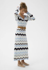 Robe en tricot avec un motif en chevrons dans des teintes de bleu, noir et marron ; comprend une ceinture beige. Portée avec des chaussures à talons beiges.