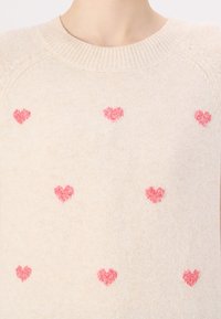 Beżowy sweter o miękkiej fakturze, okrągłym dekolcie i rozrzuconych różowych serduszkach na tkaninie.