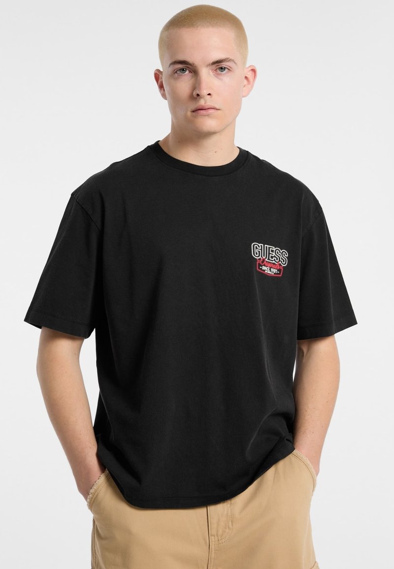 T-shirt in cotone nero con maniche corte, caratterizzato da un piccolo logo ricamato "GUESS" in rosso e bianco sul lato sinistro del petto. Vestibilità casual.