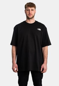 T-shirt in cotone nero con vestibilità ampia, caratterizzato da un logo bianco sul lato sinistro del petto. Collo rotondo, maniche corte e superficie del tessuto liscia.