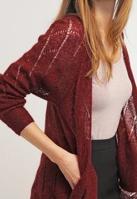 Burgundy öppenstickad cardigan med ett geometriskt mönster, ribbade ärmar och en avslappnad passform, lagd över en ljus topp och mörk kjol.