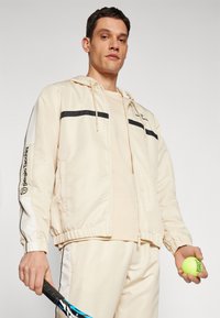 Cazadora beige con rayas negras, puños elásticos y capucha con cordón, llevada encima de una blusa beige clara. Sosteniendo una raqueta de tenis y una pelota.