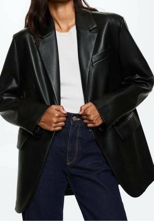 Kvinde iført oversized sort læderblazer, hvid ribbet top og højtaljede mørkeblå jeans, der holder på blazernes slag.