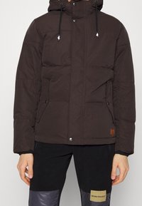 Brown padded jacket with a hood, featuring snap closures and drawstring. Made from a soft fabric with a textured finish.