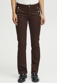 Pantalons en toile marron avec une coupe slim, dotés de boutons sur les côtés, de poches avant et d'une texture lisse. Portés avec des chaussures noires.