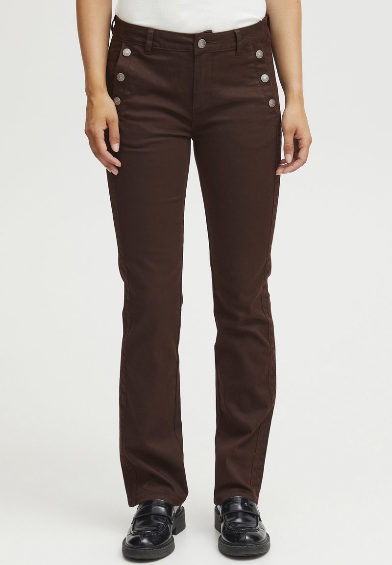 Pantalons en toile marron avec une coupe slim, dotés de boutons sur les côtés, de poches avant et d'une texture lisse. Portés avec des chaussures noires.