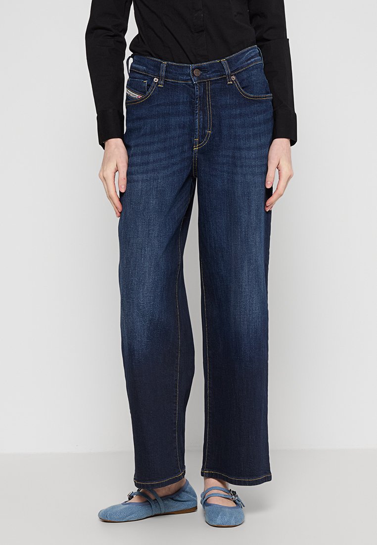 Jeans a gamba larga blu scuro con un design classico, caratterizzati da cuciture gialle e tasche frontali, abbinati a una camicia nera e ballerine blu.