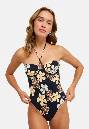 Jeune femme portant un maillot de bain noir sans bretelles avec des motifs floraux jaunes, bleus et bruns, ajustant les côtés sur un fond blanc uni.