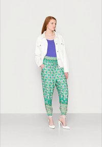 Mulher em pé, vestindo uma jaqueta branca, uma blusa roxa, calças largas com padrão verde e sapatos de salto alto brancos com tiras no tornozelo, contra um fundo liso.