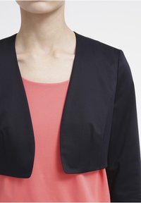 Czarna krótka marynarka o gładkiej fakturze, z krótkimi rękawami i szerokim otwartym przodem, noszona na koralowej bluzce.