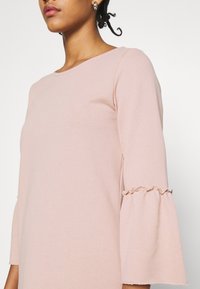 Ljusrosa långärmad topp med rundad halsringning. Har volangdetaljer vid ärmsluten och en mjuk, sl ät textur.