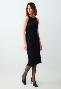 Schwarzes ärmelloses Kleid mit einem geknoteten Detail an der Taille, knielang, aus glattem Stoff, kombiniert mit schwarzen Stilettos und feinen Strumpfhosen.