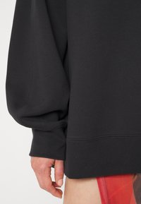 Sudadera oversized negra con dobladillo y puños acanalados. Cuenta con una suave tela texturizada. Mano visible sosteniendo la prenda.