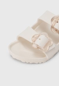 Λευκό καουτσούκ σανδάλι τύπου σλάιντ με δύο διαφανείς αγκράφες σε σχήμα λουλουδιού. Στρογγυλεμένη μύτη, υφασμένο πέλμα και ραβδωτή σόλα για καλύτερο κράτημα.