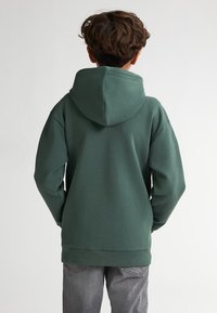 Tmavě zelená bavlněná mikina s velkou kapucí, spadlými rameny a žebrovanými manžetami, nošená přes šedé džíny. Hladká textura, volný střih.