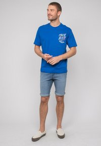 T-shirt en coton bleu avec un design graphique sur la poitrine gauche et des manches retroussées, associé à un short en denim clair et des chaussures à enfiler.