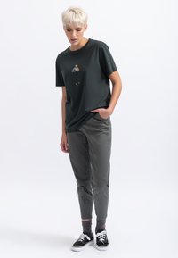 Donkergroen katoenen t-shirt met een kleine kleurrijke afbeelding op de voorkant, gecombineerd met lichtgrijze tapered broek en zwarte sneakers.