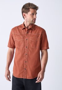 Short-sleeve, rust-colored shirt in textured fabric, featuring two chest pockets, black buttons, and contrasting stitching. Casual fit.